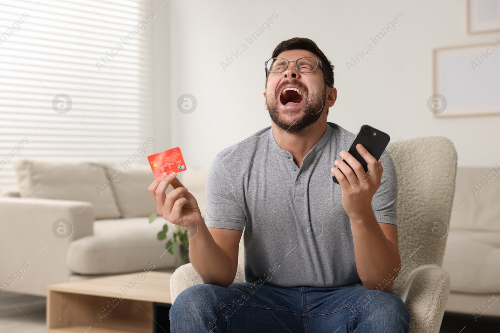 Photo of Emotional man with credit card and smartphone in armchair at home. Be careful - fraud