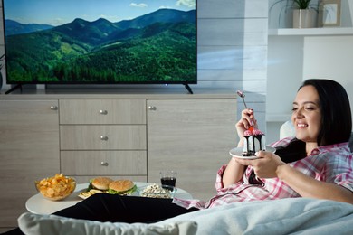 Photo of Happy overweight woman eating cake while watching TV on sofa at home