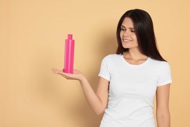 Beautiful young woman holding bottle of shampoo on beige background