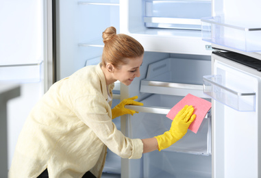 Woman in rubber gloves cleaning empty refrigerator with rag at home