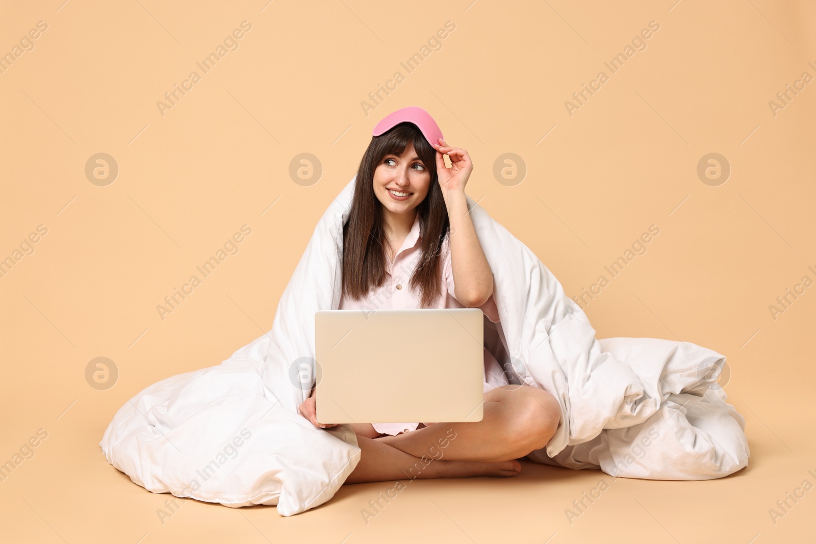 Photo of Happy woman with pyjama and blanket holding laptop on beige background