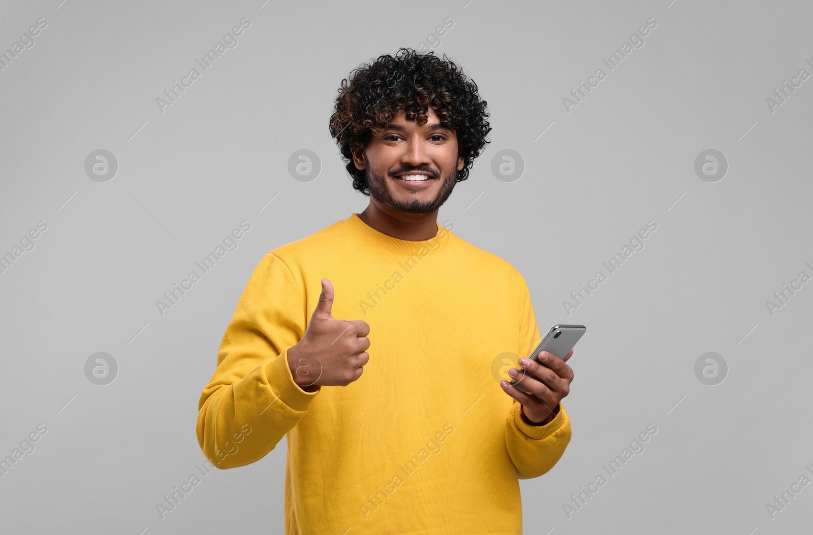 Photo of Handsome smiling man using smartphone and showing thumbs up on light grey background