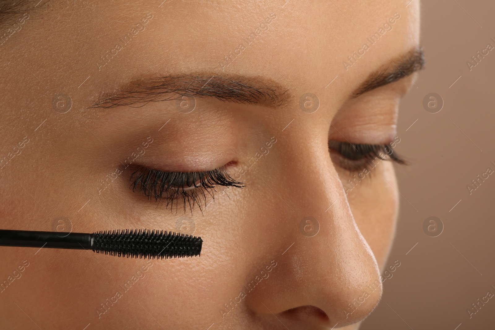 Photo of Woman applying mascara onto eyelashes against light brown background, closeup