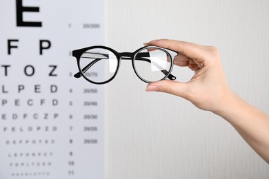 Photo of Woman holding glasses against eye chart on light background, closeup. Ophthalmologist prescription