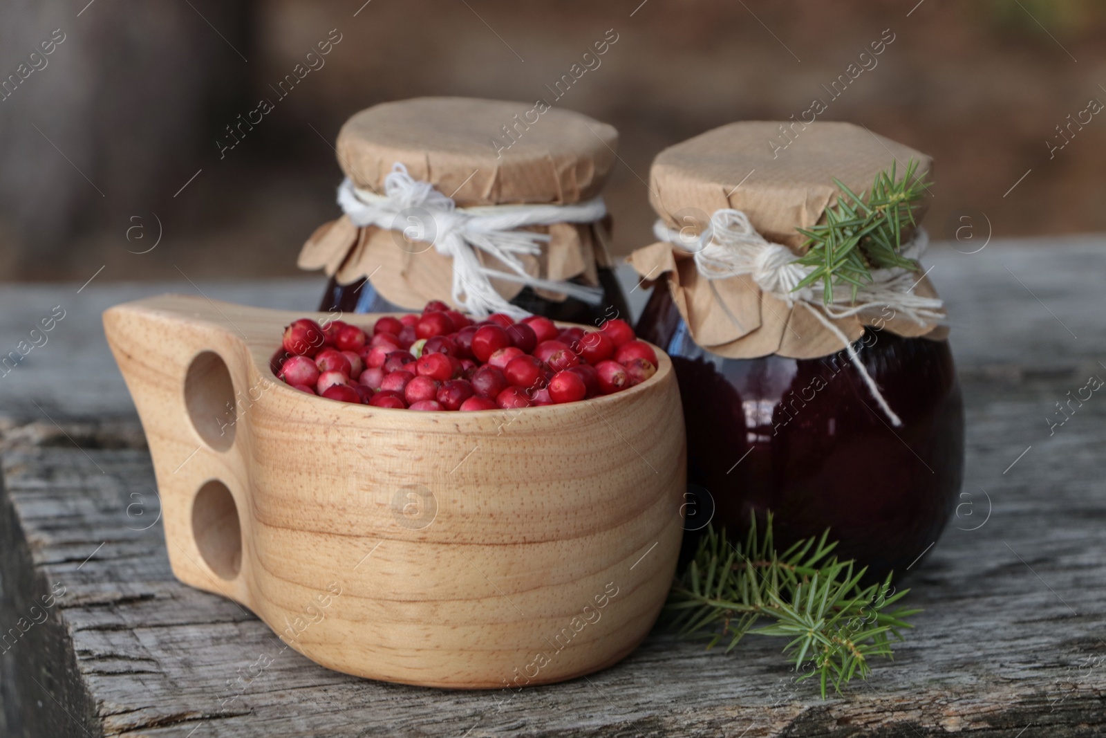 Photo of Tasty lingonberry jam in jars and cup with red berries on wooden table outdoors