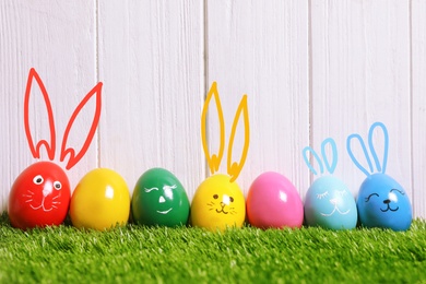 Image of Several eggs with drawn faces and ears as Easter bunnies among others on green grass against white wooden background