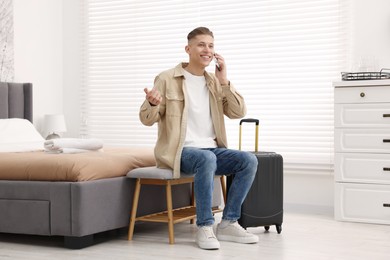 Photo of Smiling guest with suitcase talking on smartphone in stylish hotel room