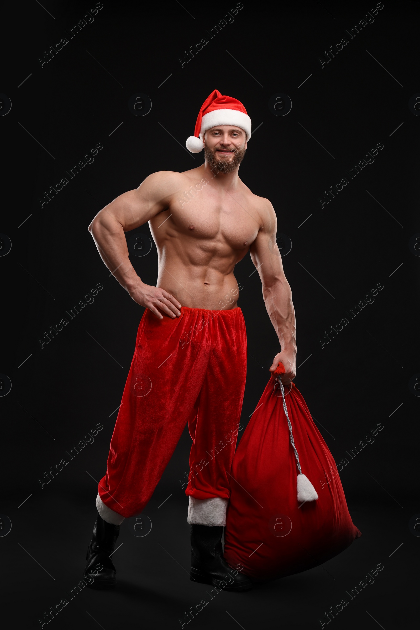 Photo of Muscular young man in Santa hat holding bag with presents on black background