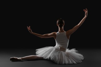 Photo of Young ballerina sitting on split against black background, back view