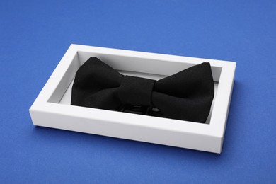 Stylish black bow tie in box on blue background, closeup