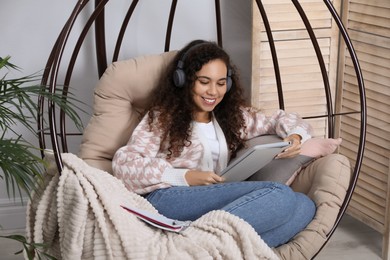 Photo of African American woman with headphones and tablet studying in egg chair at home. Distance learning