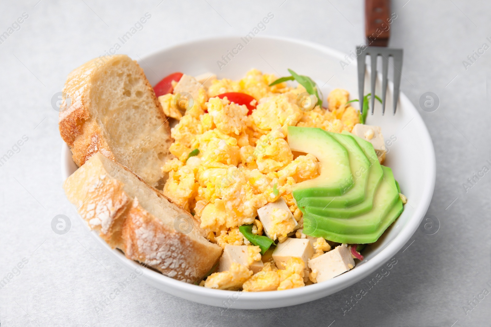 Photo of Bowl with delicious scrambled eggs, tofu, avocado and slices of baguette on white table, closeup