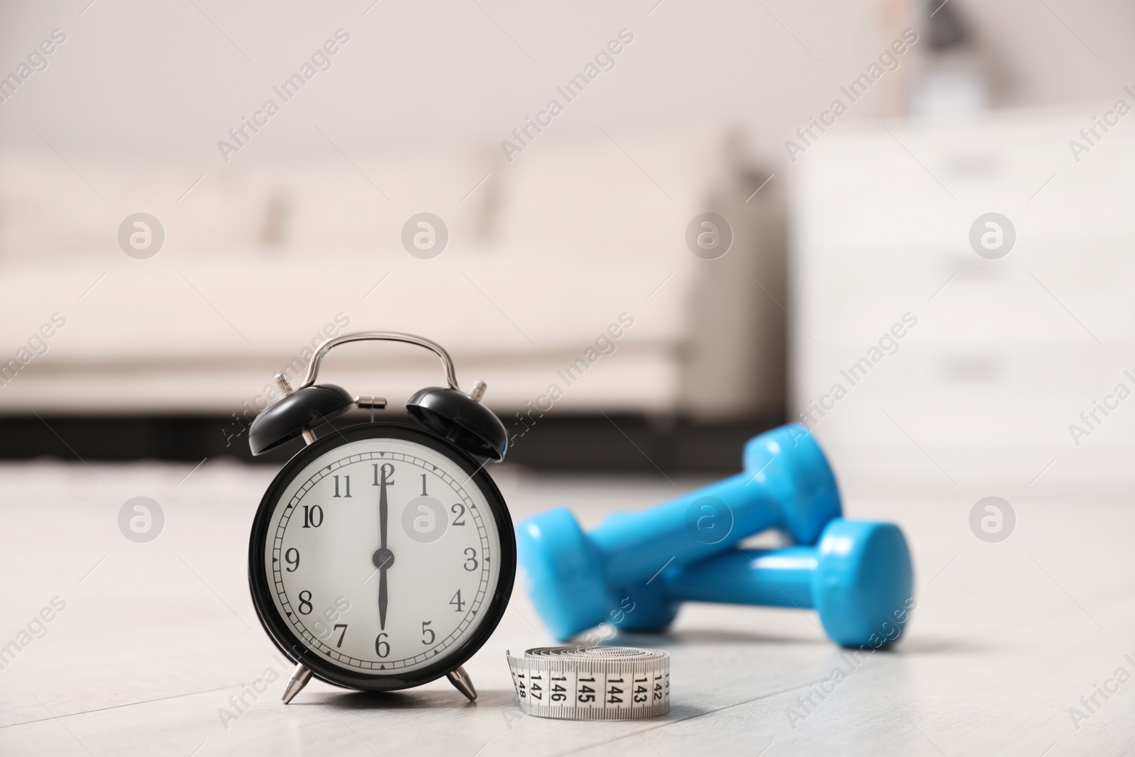 Photo of Alarm clock, measuring tape and dumbbells on wooden floor indoors. Morning exercise