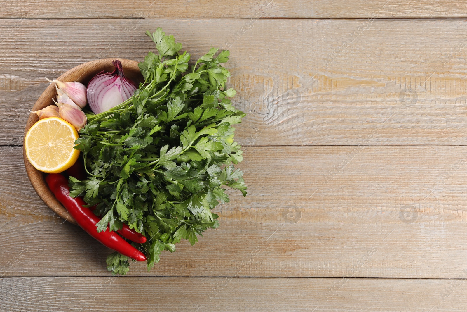 Photo of Bowl with fresh green parsley, chili peppers, lemon, onion and garlic on wooden table, top view. Space for text