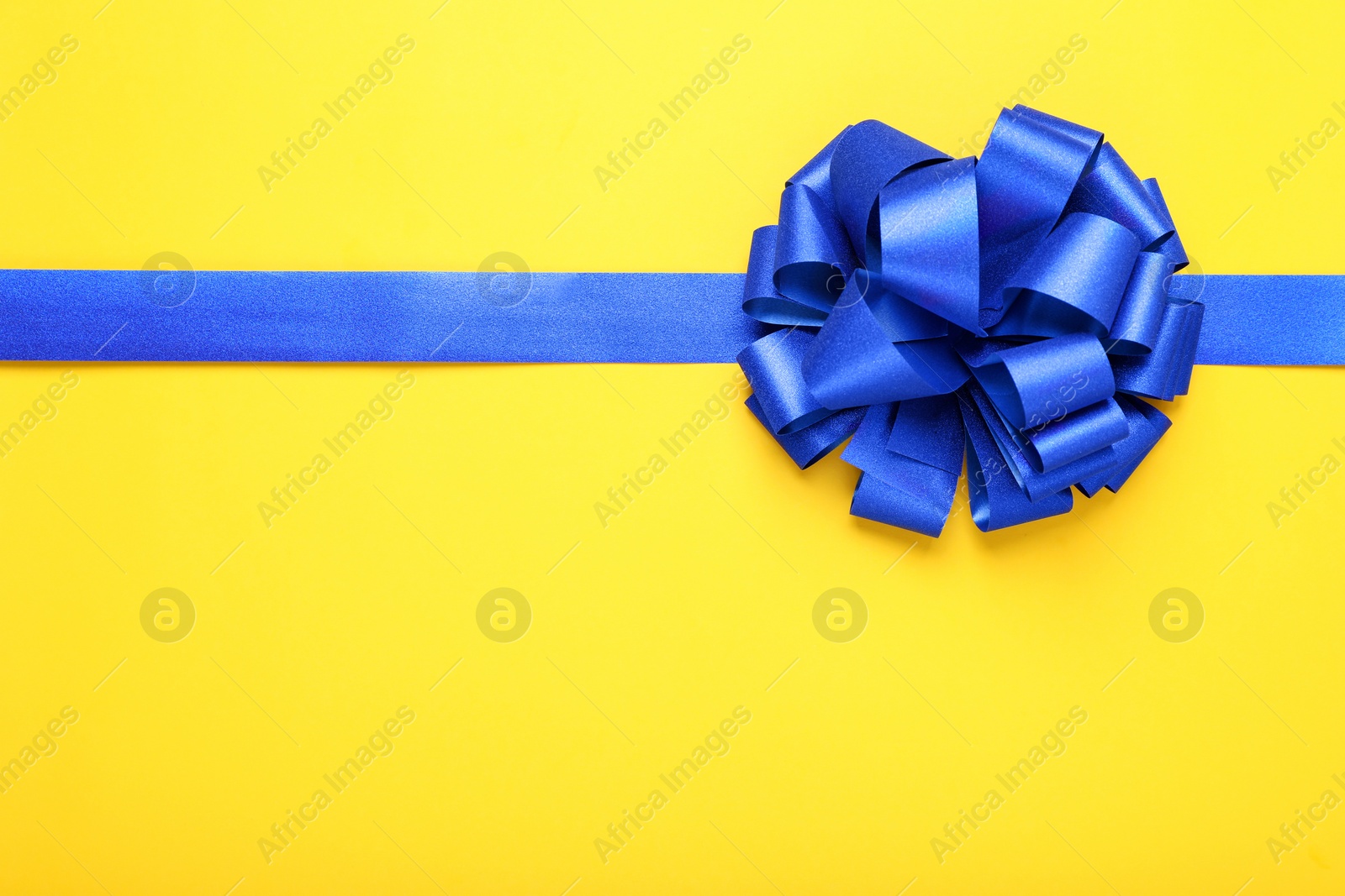 Photo of Blue ribbon with bow on yellow background, top view. Space for text