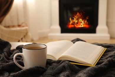 Photo of Cup of hot tea and book on knitted plaid near fireplace at home. Cozy atmosphere
