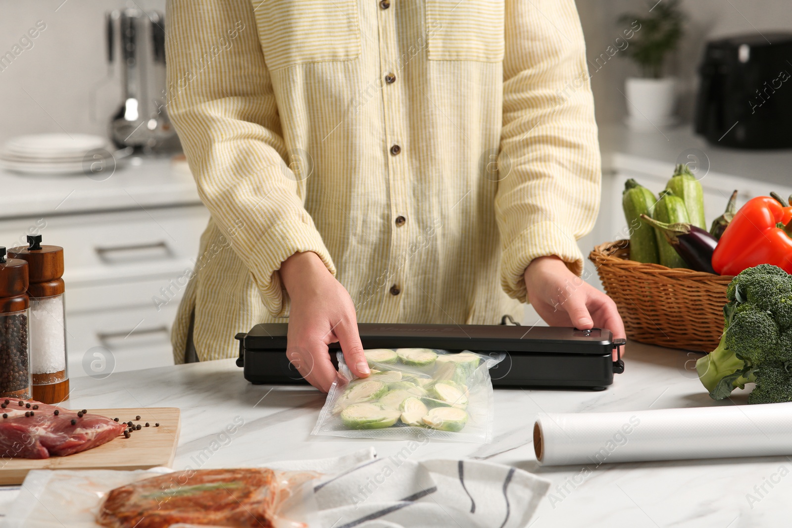 Photo of Woman using sealer for vacuum packing with plastic bag of zucchini at white table in kitchen, closeup