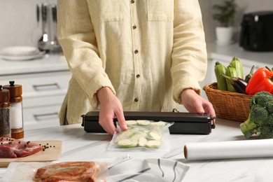 Photo of Woman using sealer for vacuum packing with plastic bagzucchini at white table in kitchen, closeup