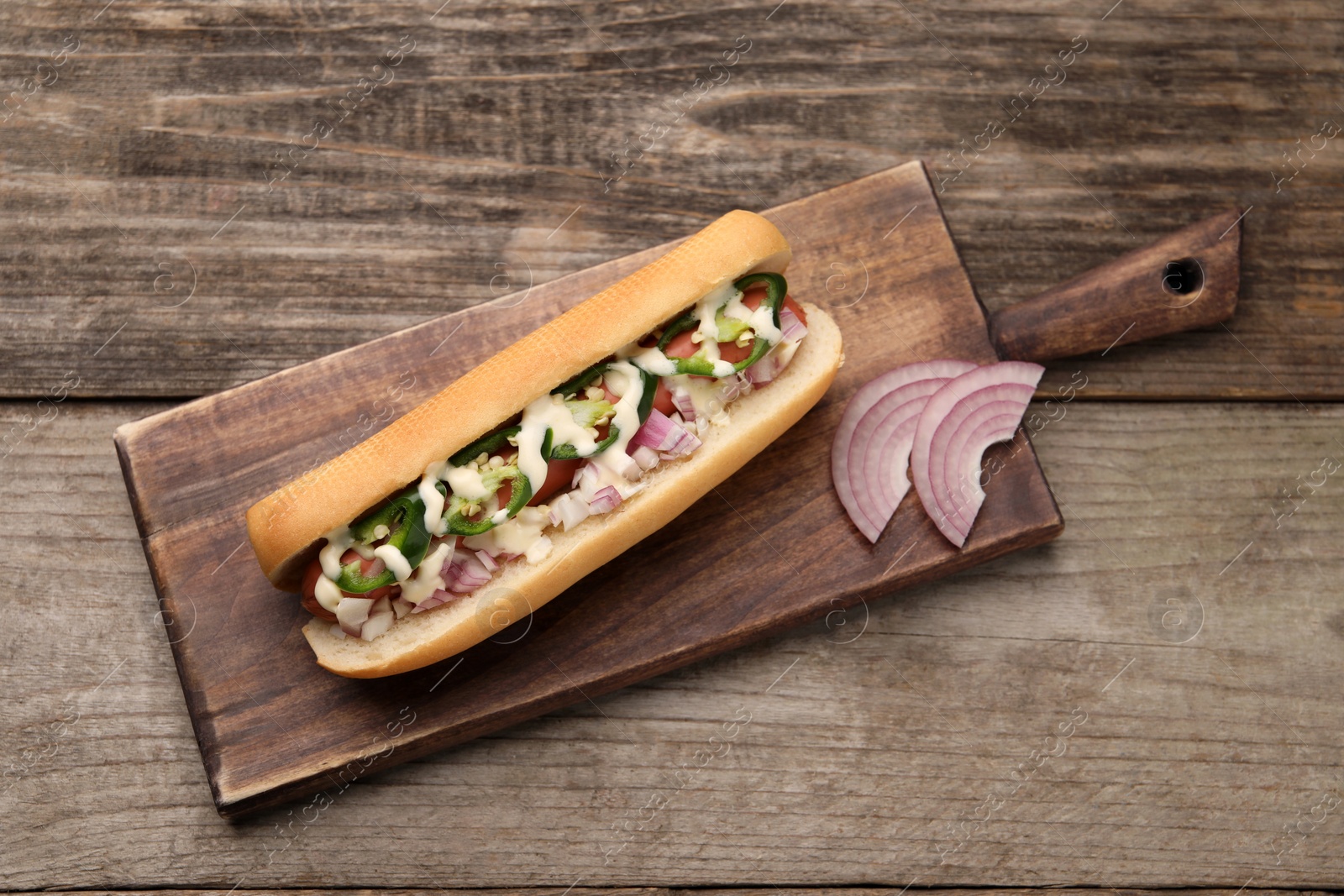 Photo of Delicious hot dog with onion, chili pepper and sauce on wooden table, top view