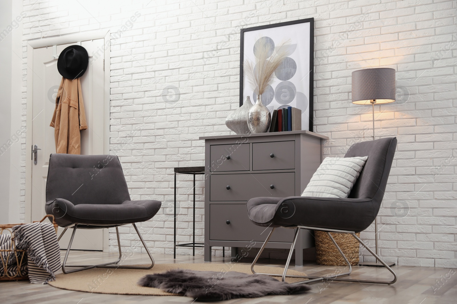 Photo of Stylish room interior with grey chest of drawers and chairs near white brick wall