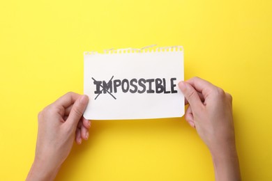 Photo of Motivation concept. Woman holding paper with changed word from Impossible into Possible by crossing over letters I and M on yellow background, top view