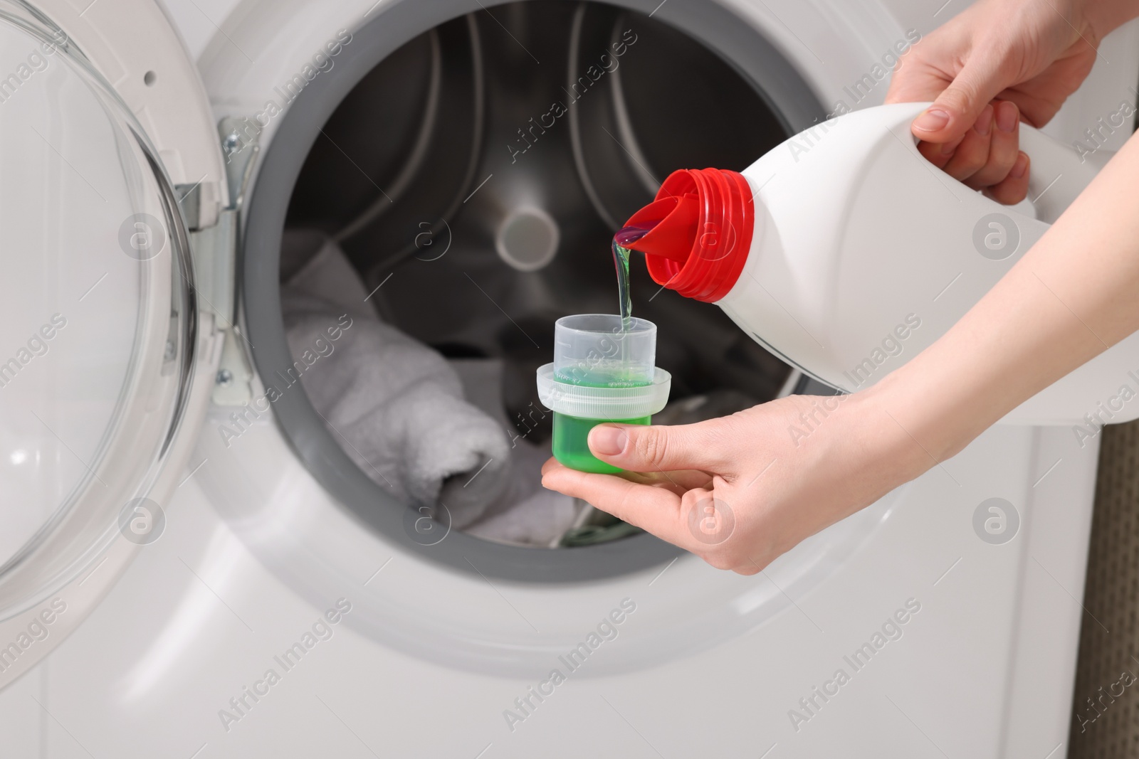 Woman pouring fabric softener from bottle into cap near washing machine, closeup Photo of Woman pouring fabric softener from bottle into cap near washing machine, closeup