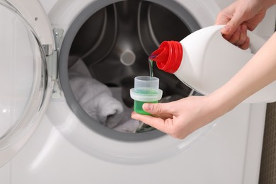 Photo of Woman pouring fabric softener from bottle into cap near washing machine, closeup