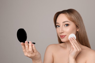 Beautiful young woman applying face powder with puff applicator on grey background