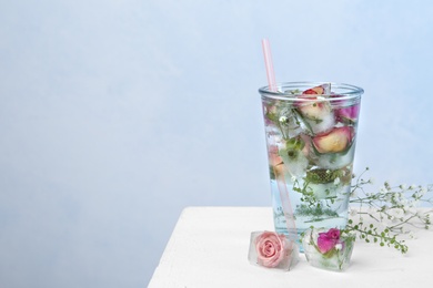 Photo of Glass of water with ice cubes and flowers on table against color background, space for text