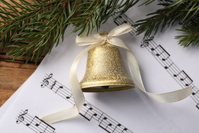 Photo of Golden shiny bell with bow, music sheet and fir branches on wooden table, flat lay. Christmas decoration