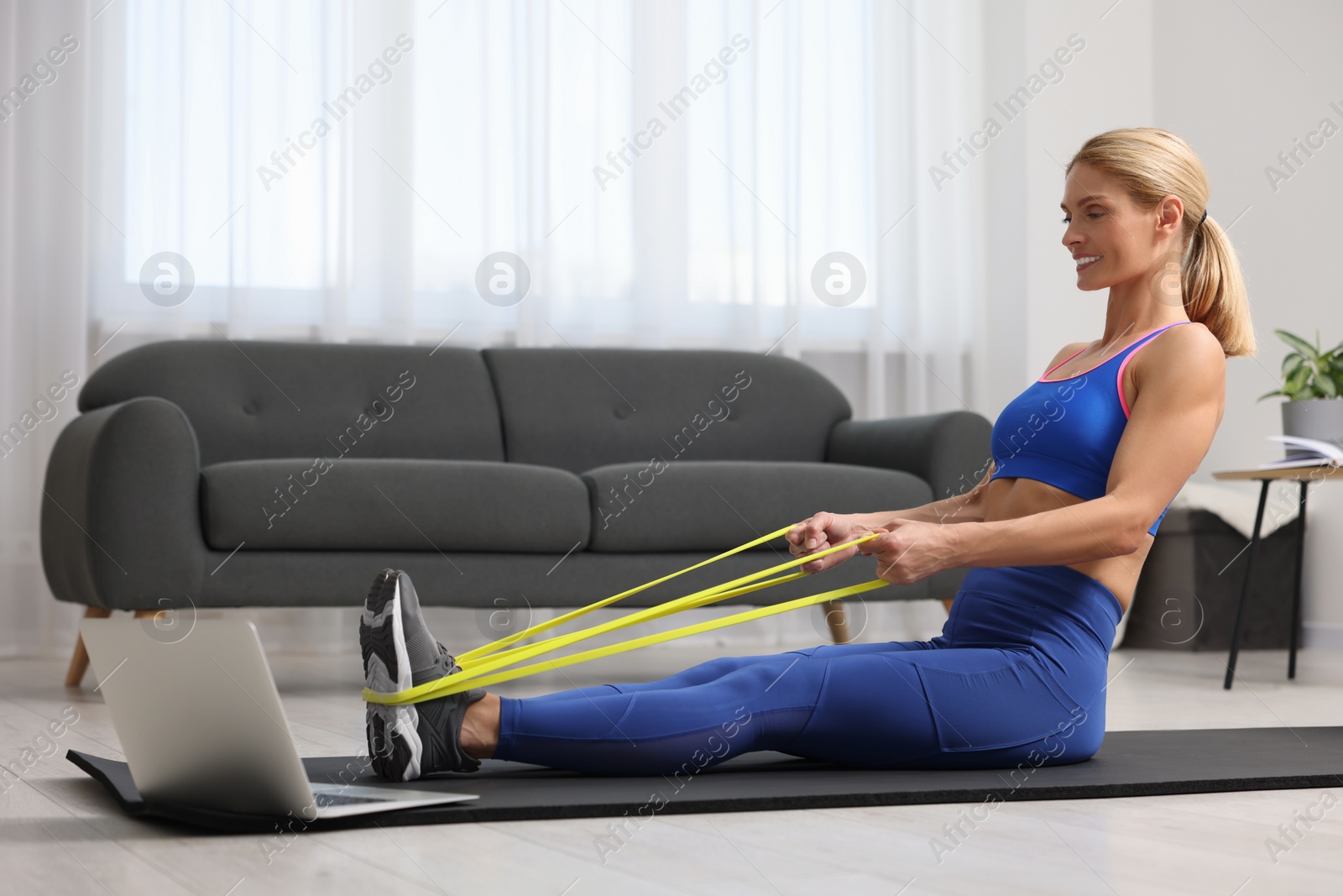 Photo of Fit woman doing exercise with fitness elastic band near laptop on mat at home