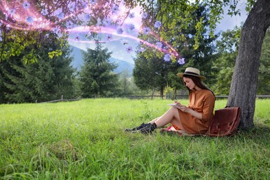 Beautiful young woman drawing with pencil in notepad near tree on green grass. Flowers flying near her symbolizing state of mindfulness