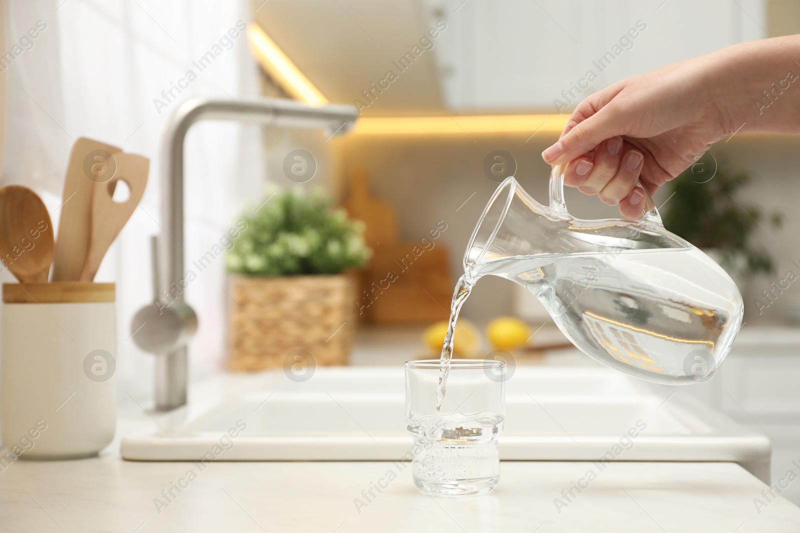 Photo of Woman pouring water from jug into glass at white table in kitchen, closeup. Space for text