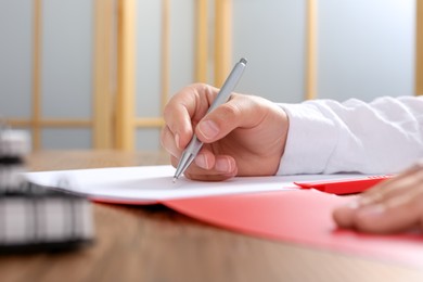 Woman writing on sheet of paper in red folder at wooden table in office, closeup