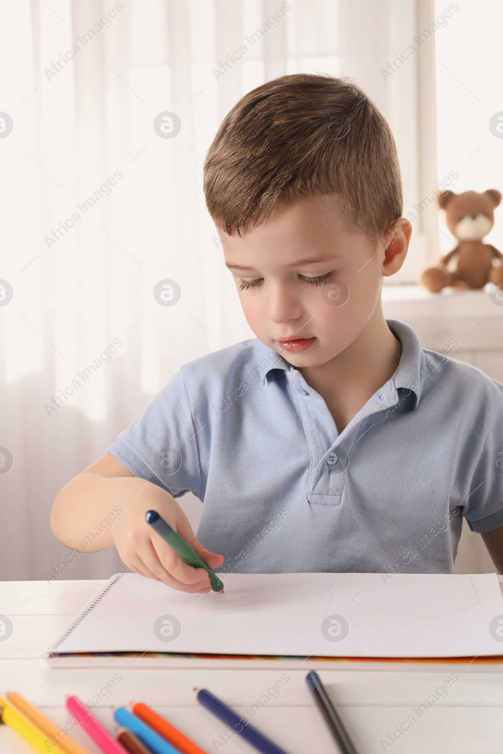 Photo of Cute little boy drawing with pencil at white table in room. Child`s art