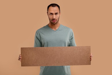 Upset man holding blank cardboard banner on beige background, space for text
