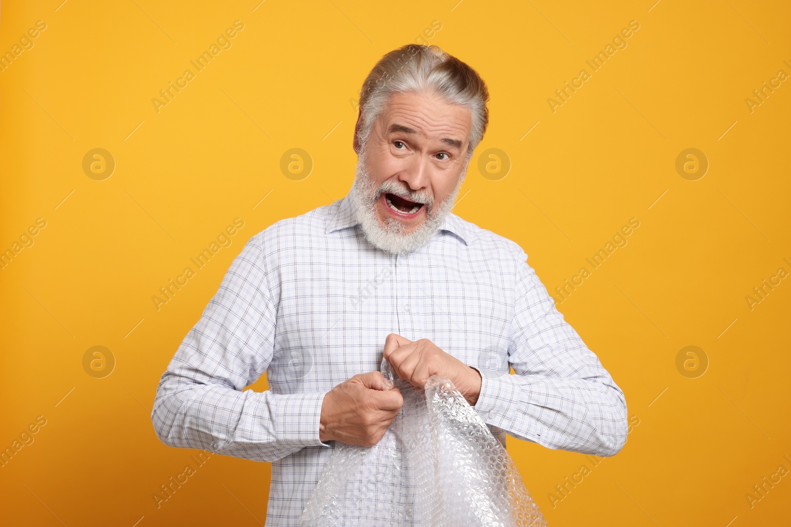 Photo of Emotional senior man popping bubble wrap on yellow background. Stress relief