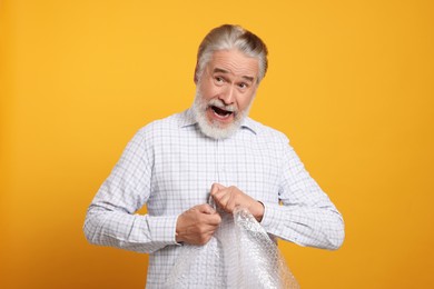 Emotional senior man popping bubble wrap on yellow background. Stress relief