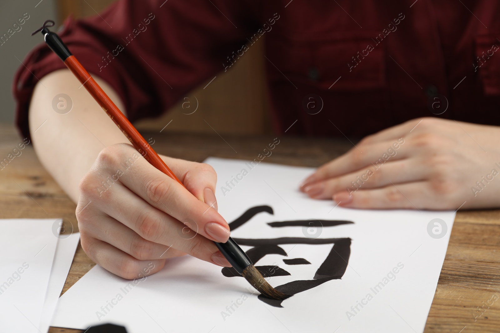 Photo of Calligraphy. Woman with brush writing hieroglyphs on paper at wooden table, closeup