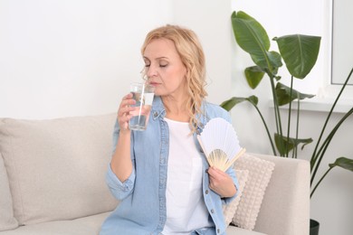 Photo of Woman with glass of water and hand fan on sofa at home. Hormonal disorders