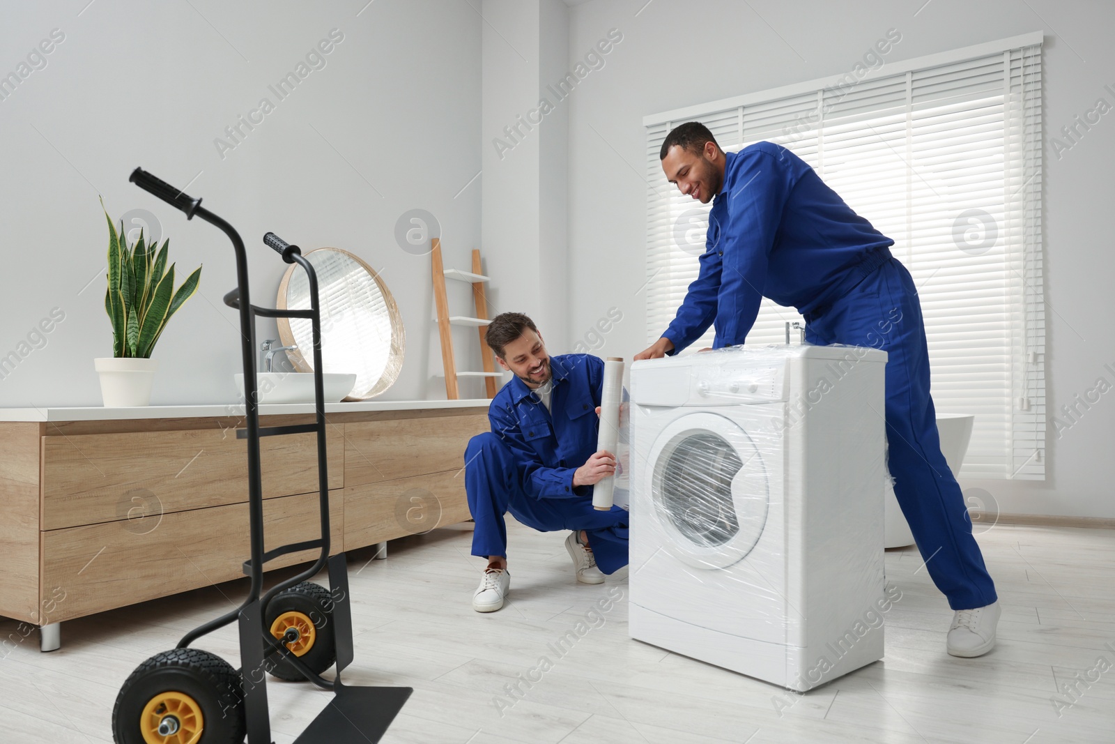 Photo of Male movers with stretch film wrapping washing machine in bathroom. New house