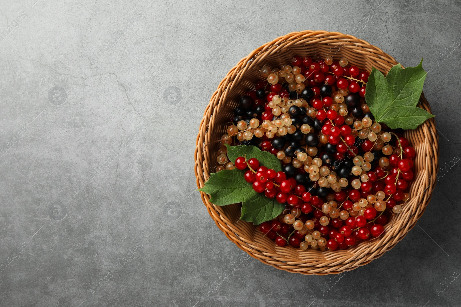 Photo of Different fresh ripe currants and green leaves in wicker basket on light grey table, top view. Space for text