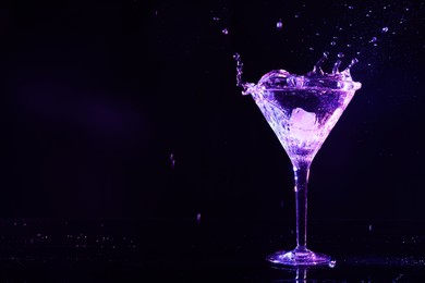 Photo of Glass of martini with ice and splashes in neon lights on dark background. Space for text
