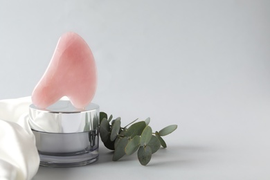 Photo of Rose quartz gua sha tool, jar of cream and eucalyptus branches on grey background, space for text