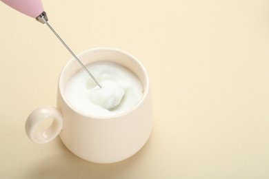 Whisking milk in cup with mini mixer (milk frother) on beige background. Space for text