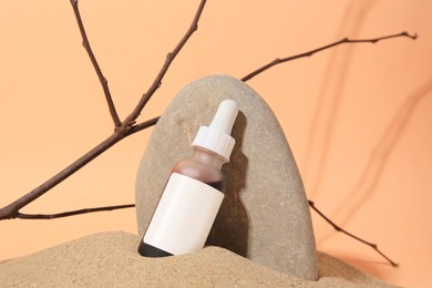 Photo of Bottle with serum, branches and stone on sand against orange background. Cosmetic product
