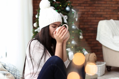 Beautiful young woman in hat with cup of drink at home. Christmas celebration