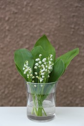 Photo of Beautiful lily of the valley flowers in glass vase on white table near brown wall