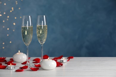 Glasses of champagne, candles and rose petals on white wooden table against blurred festive lights, space for text