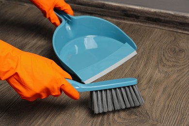Photo of Woman in gloves sweeping wooden floor with plastic whisk broom and dustpan indoors, closeup